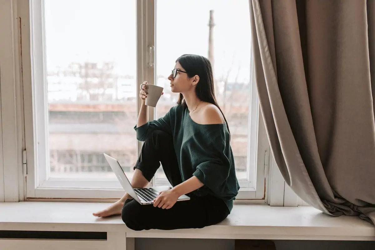 Pensive woman with laptop drinking hot beverage on windowsill